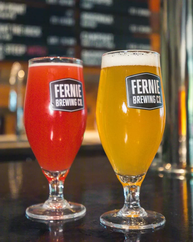 Two tulip glasses of beer on a bar counter, one red and one yellow, both labeled Fernie Brewing Co. A blurred menu board is in the background—a perfect stop for your Fernie guide or list of things to do in Fernie.