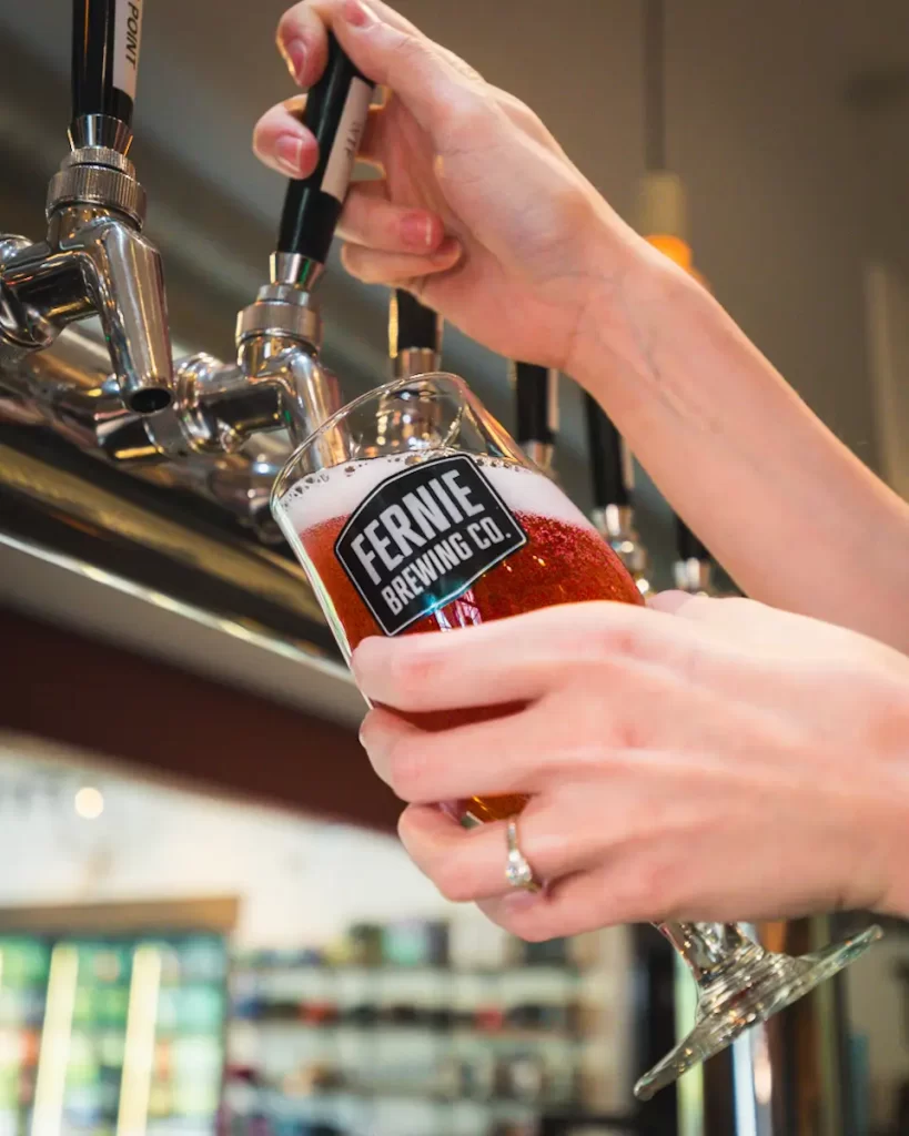 A person fills a Fernie Brewing Co. glass with amber beer from a tap at a bar, showcasing one of the top things to do in Fernie for beer lovers and travelers exploring local Fernie attractions.