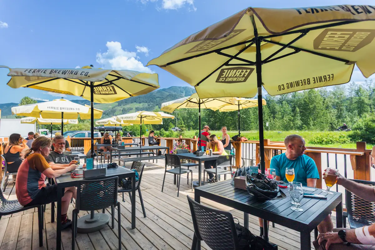 People sit at tables under large yellow umbrellas on a wooden patio, enjoying drinks and conversation on a sunny day. Surrounded by green trees and mountains, it’s the perfect spot to relax—one of many Fernie attractions worth adding to your itinerary.