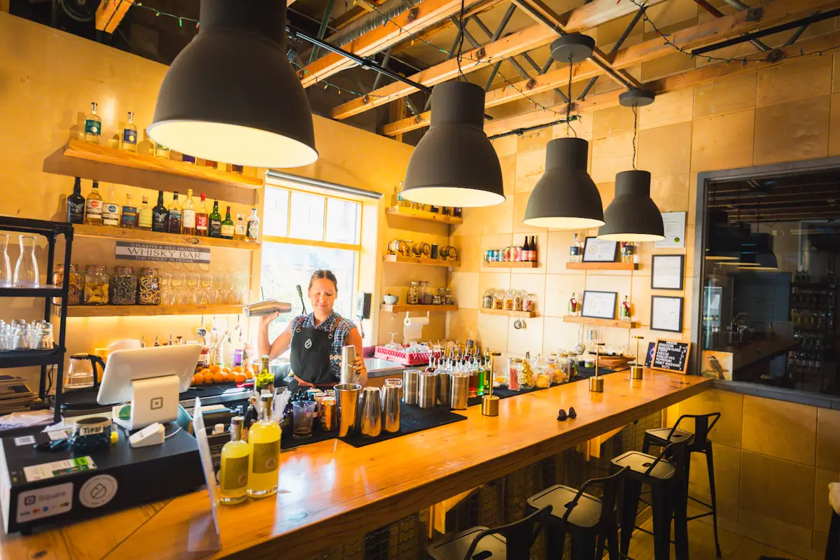 A bartender stands behind a wooden bar, shaking a drink. Bottles, glassware, and mixers are arranged on shelves. Large black pendant lights hang from the ceiling in a warmly lit, modern bar—a must-visit spot featured in any Fernie guide.