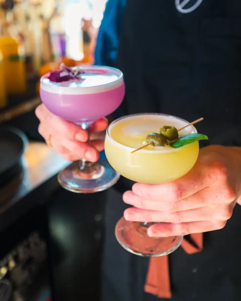 A person in a dark apron holds two vibrant cocktails—one purple with a flower garnish, the other yellow with green—served in coupe glasses. Discover unique flavors like these among the many things to do in Fernie.