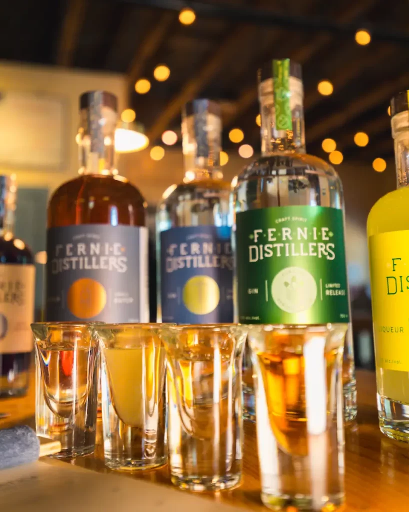 Four bottles of Fernie Distillers spirits are lined up on a wooden table, each with a different colored label. Shot glasses sit ready in front, making this tasting a must-try on any Fernie guide or list of things to do in Fernie. Warm string lights glow behind.