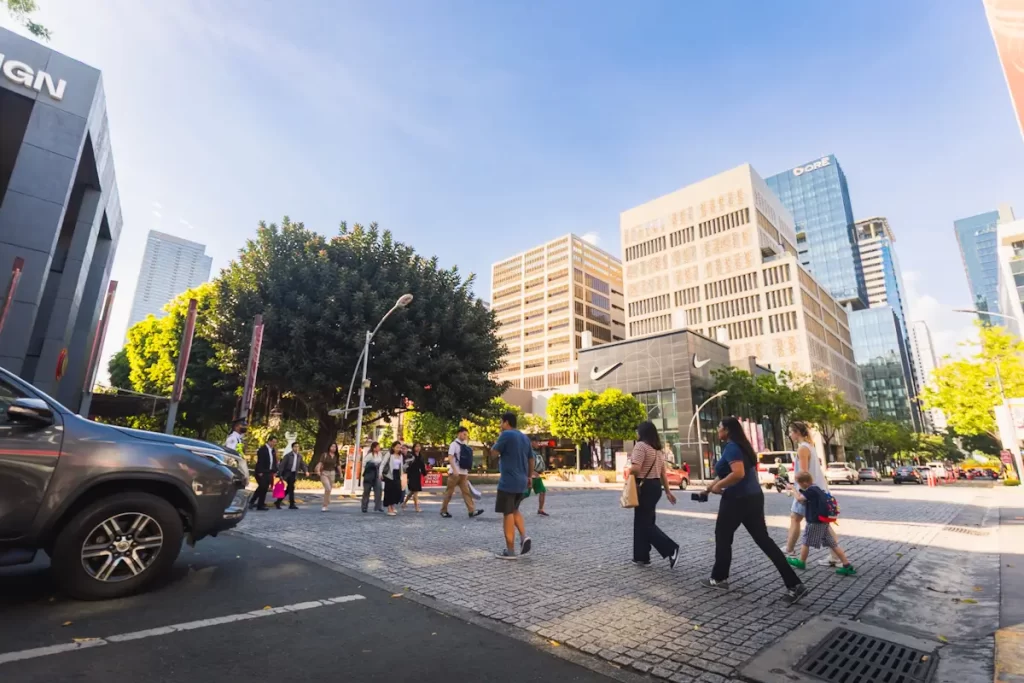 People cross a city street at a pedestrian crosswalk on a sunny day, surrounded by buildings, trees, and parked vehicles—capturing the vibrant atmosphere near Manila tourist spots under a clear blue sky.