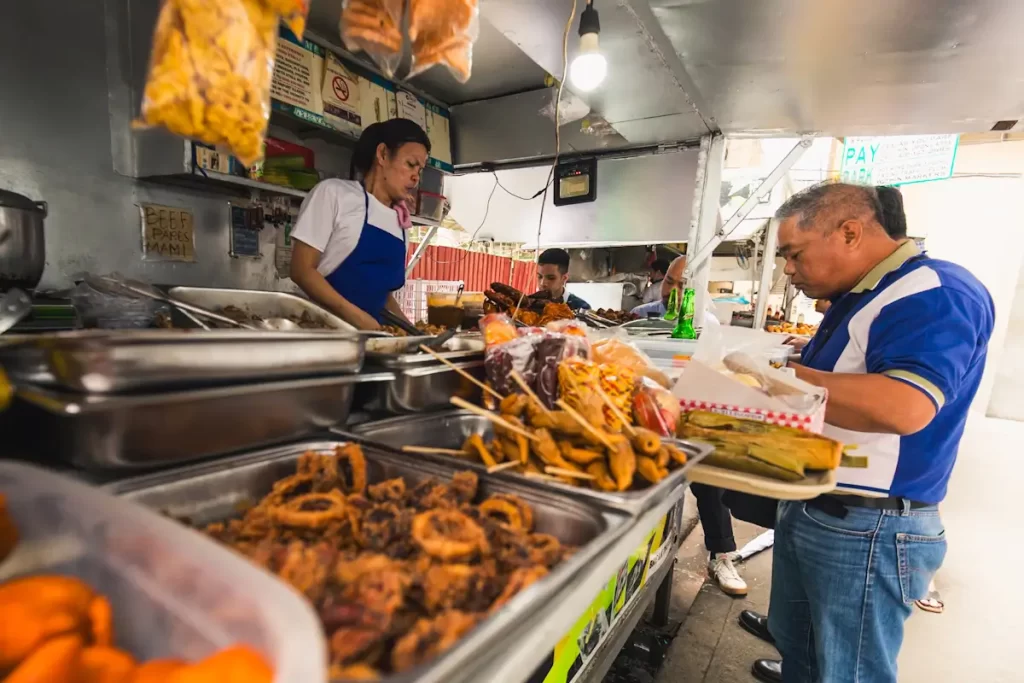 A man in a blue shirt selects food from a variety of fried items and snacks at an outdoor food stall, one of the lively Manila tourist spots, while a woman behind the counter prepares to assist customers. Other people are eating in the background.