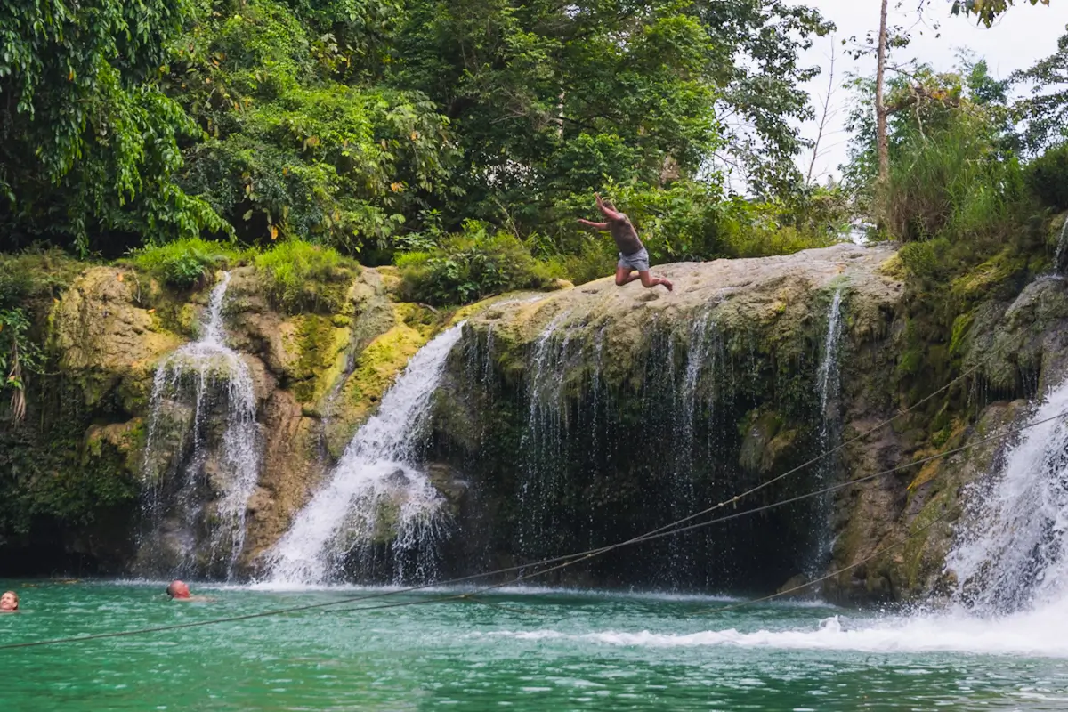 A person is mid-jump off a rocky ledge above Pangas Falls, a stunning tourist attraction where two waterfalls cascade into a green pool surrounded by dense foliage and trees.