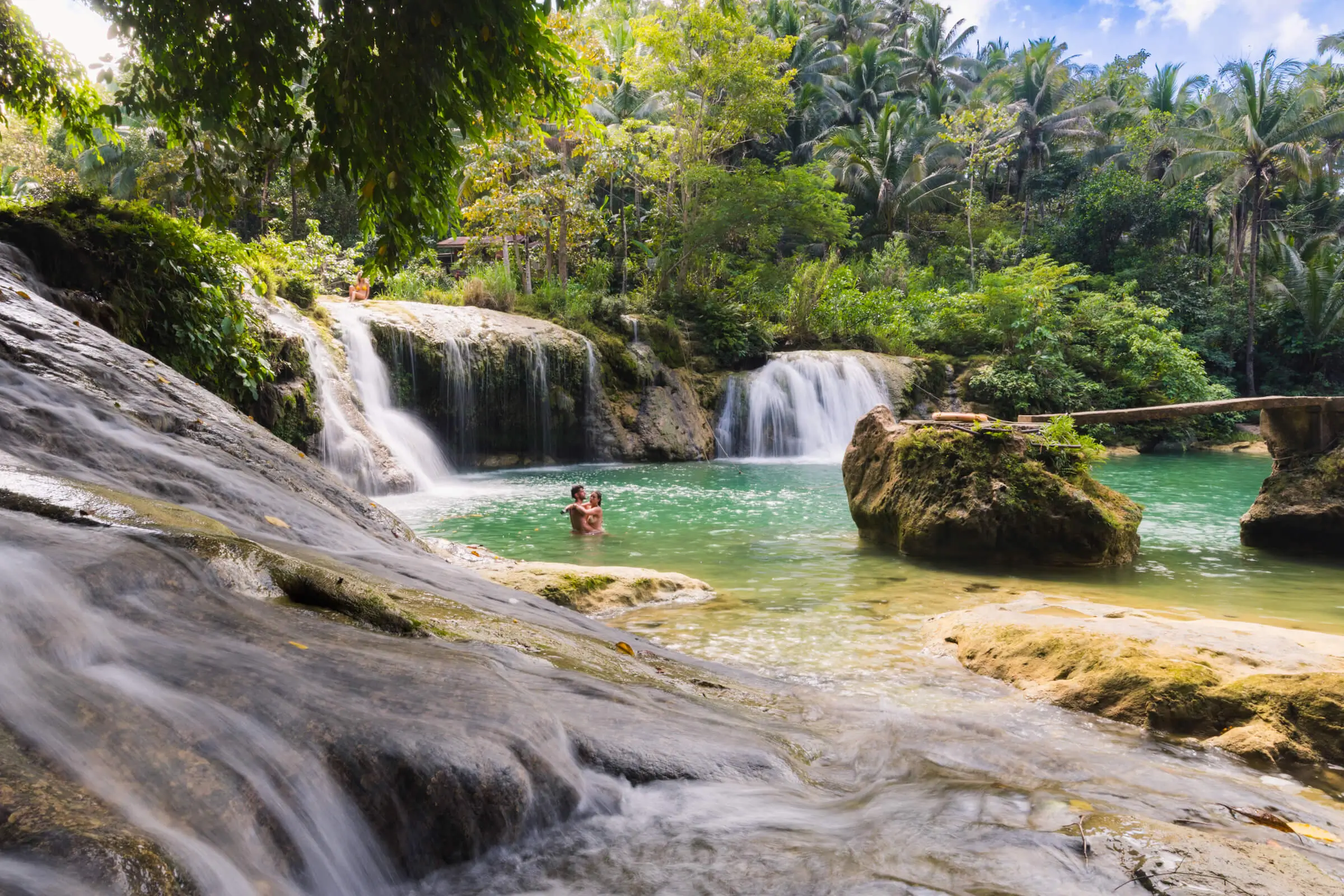 A person stands in the clear pool beneath two small waterfalls at Pangas Falls, a hidden tourist attraction surrounded by lush green trees and dense forest. Sunlight filters through the foliage as water flows over smooth stones in the foreground.