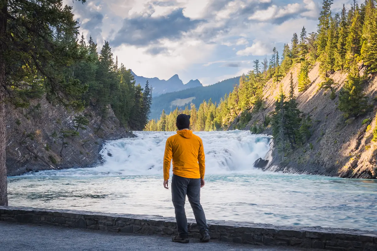 A person in a yellow jacket stands on a stone path facing a river and waterfall, surrounded by pine trees and mountains under a partly cloudy sky—capturing the essence of Banff for any Banff Guide or list of Things to Do in Banff.
