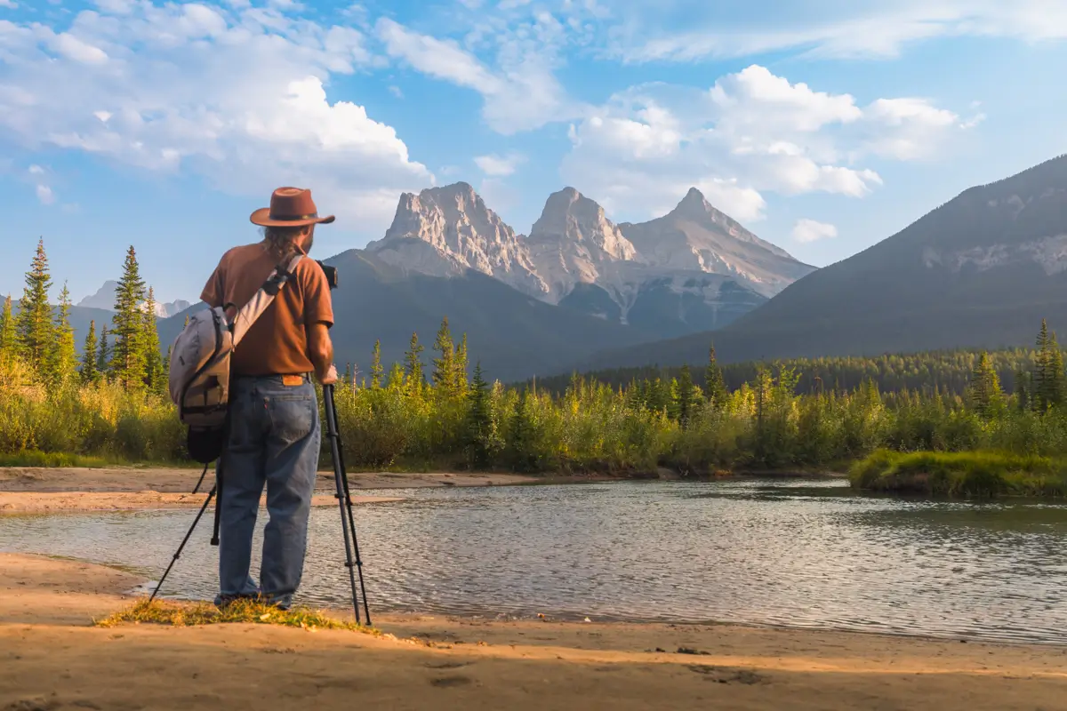 A person wearing a hat stands by the riverbank in Canmore with a camera and tripod, gazing toward a range of mountains under a partly cloudy sky, surrounded by trees and greenery.