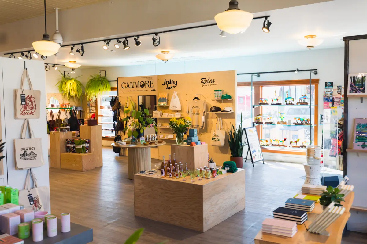 A brightly lit Canmore gift shop with wooden displays holds bags, bottles, books, mugs, plants, and souvenirs. Shelves by the window showcase colorful figurines. Signs and local artwork decorate the walls.