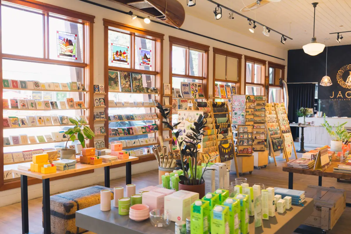 A well-lit Canmore gift shop with shelves of greeting cards, tables displaying skincare products and candles, large windows, stained glass panels, and indoor plants arranged around the space.