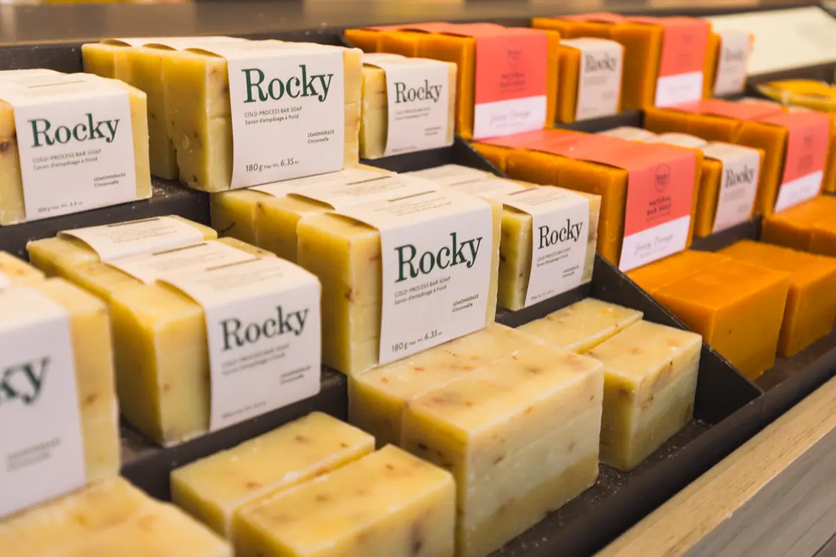 Several rows of rectangular handmade soap bars, some with Rocky labels and others with plain orange wrappers, are displayed on black trays in a Canmore retail setting.