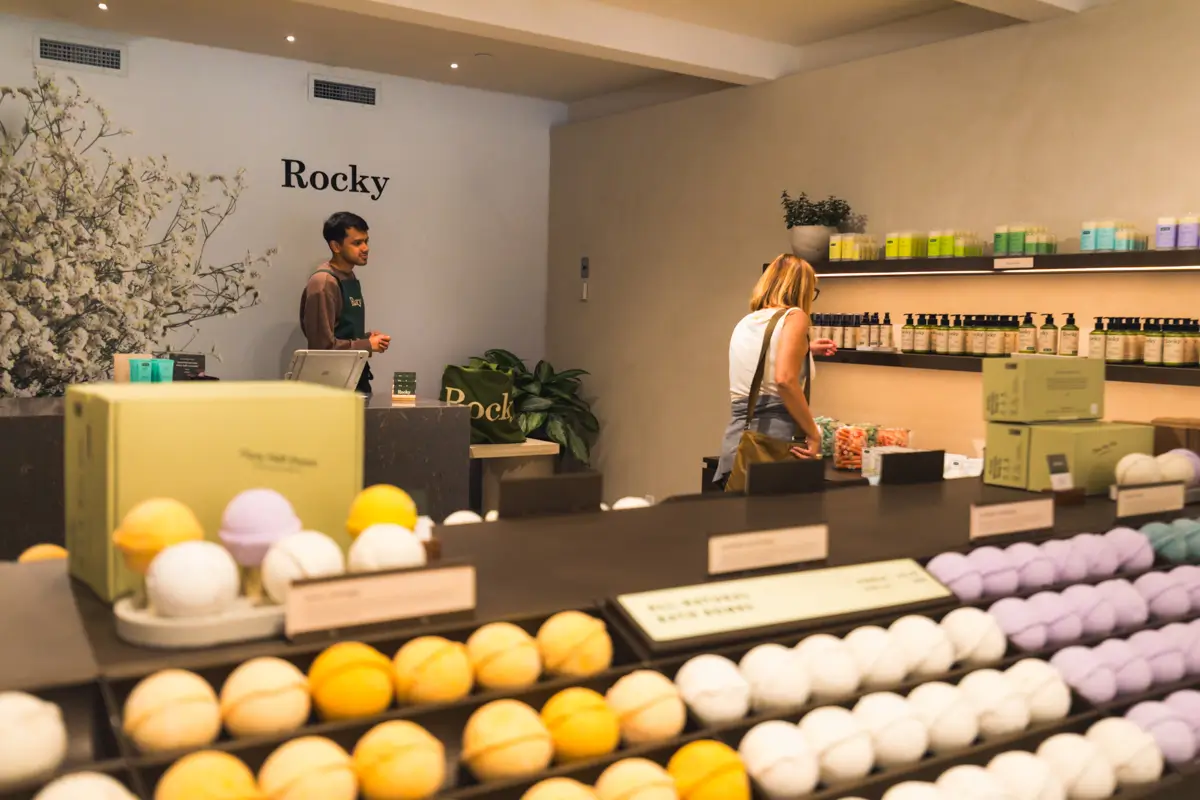A store in Canmore displays assorted bath bombs in rows on dark shelves. A woman browses products along the wall, while a staff member stands behind the counter. Shelves of colorful bottles and a decorative plant are visible.