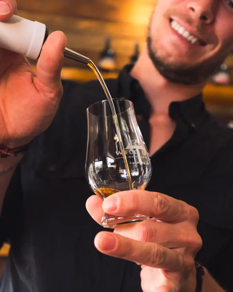 A person in a black shirt pours a small amount of brown liquid, likely whiskey, from a bottle into a whiskey tasting glass while smiling, enjoying the cozy ambiance of Canmore.