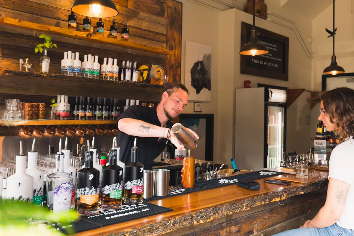 A bartender pours a drink from a shaker into a glass at a wooden Canmore bar, while a person sits across from him. Various bottles and bar tools are visible on the counter and shelves behind the bartender.