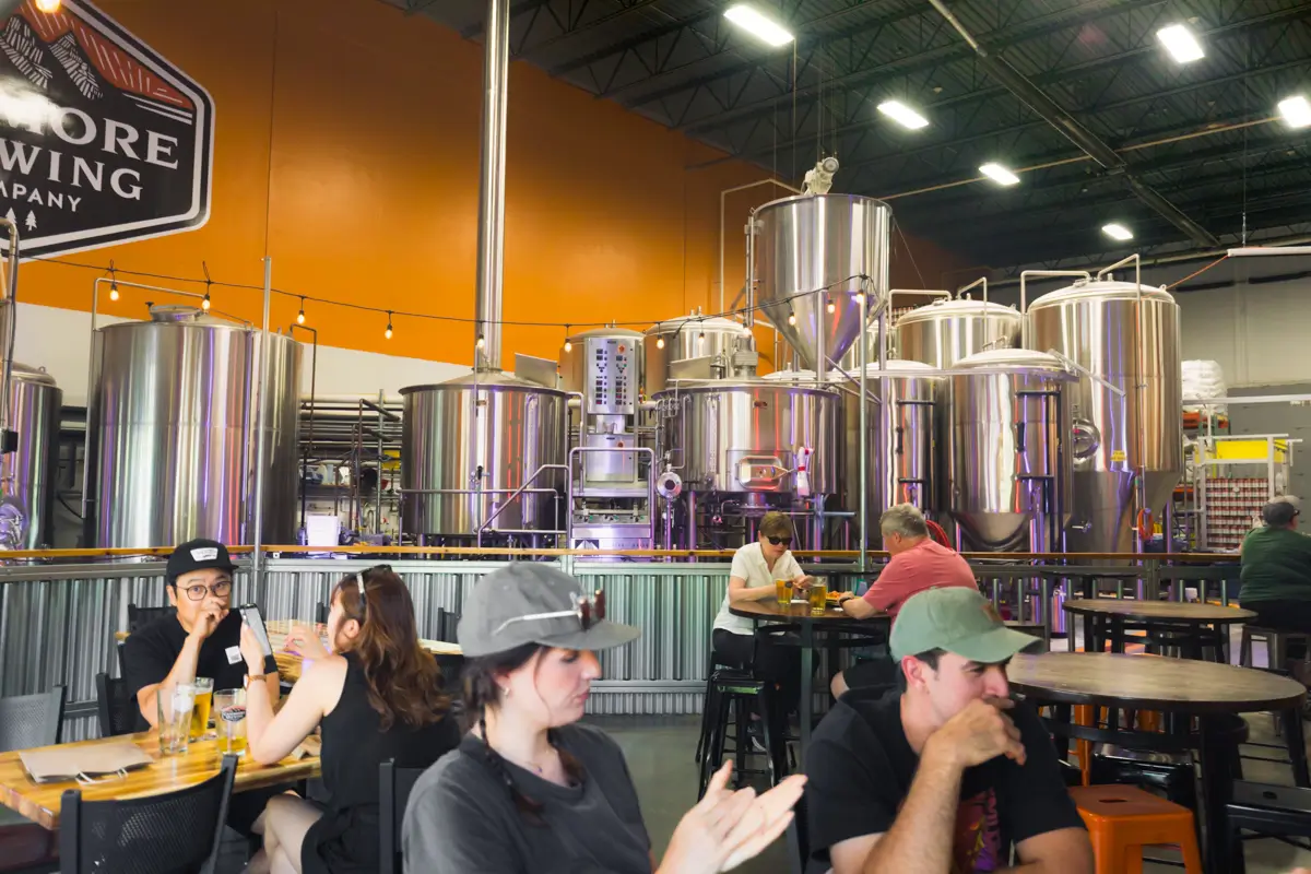 People sit at tables inside a Canmore brewery taproom, with large stainless steel brewing tanks in the background; the industrial setting features high ceilings and an orange accent wall.