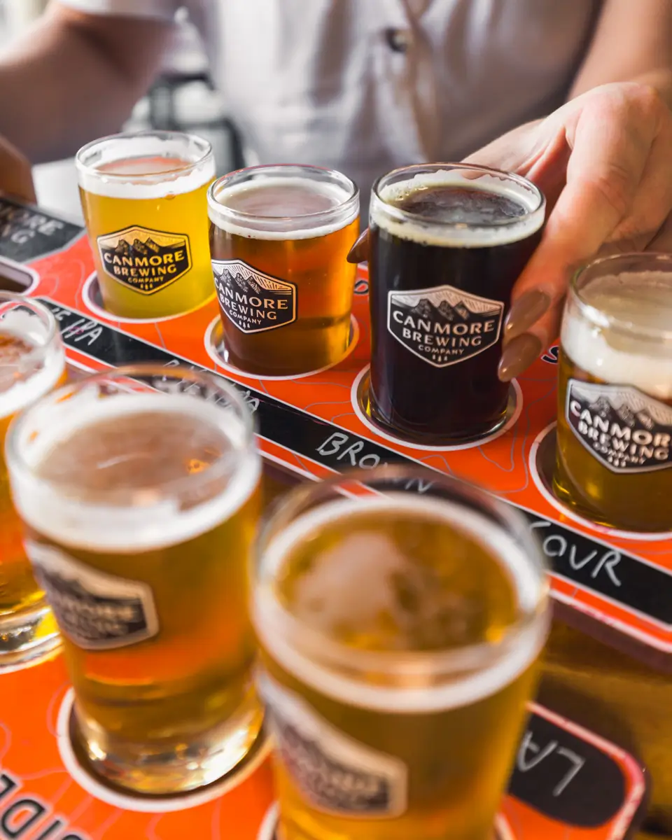 A person holds a small glass of dark beer from a flight of assorted craft beers at Canmore Brewing. The red tray, labeled “Canmore Brewing,” displays glasses with different beer colors arranged nearby.