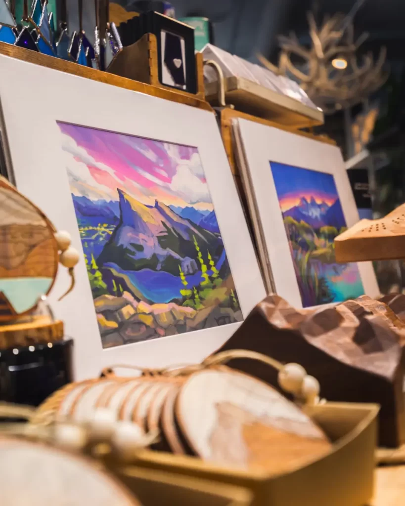 Two colorful landscape paintings of mountains and trees, inspired by Canmore, are displayed in white mats on a table surrounded by various wooden crafts and small decorative objects.
