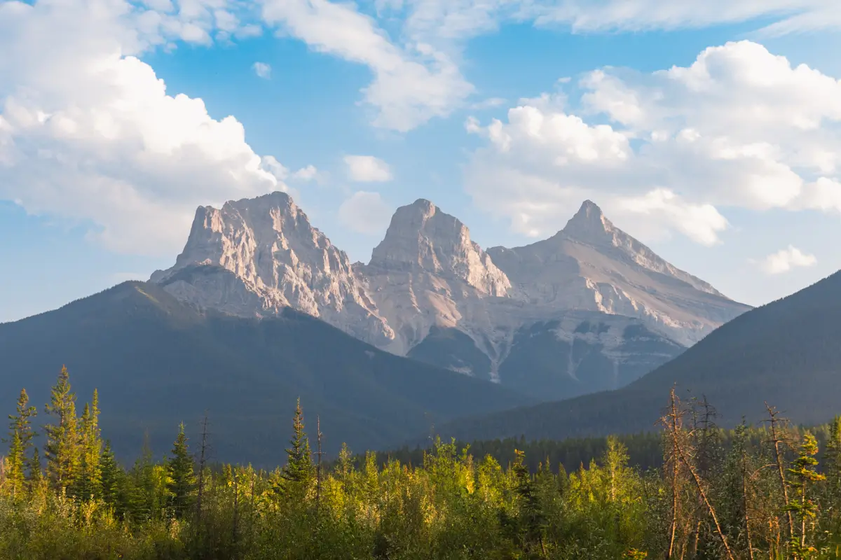 Three rocky mountain peaks rise above a forest of green trees near Canmore, set against a blue sky with scattered white clouds. The lighting suggests late afternoon or early evening.
