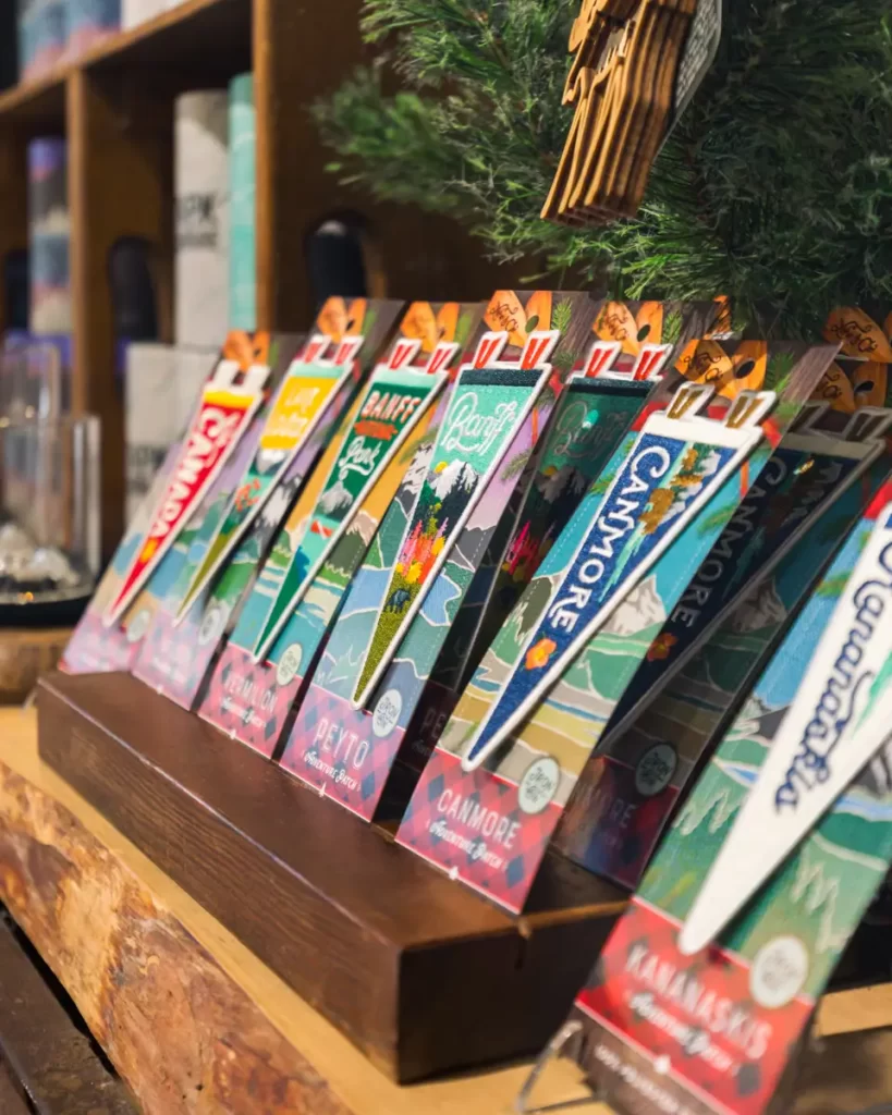 Several colorful, illustrated pennant-shaped bookmarks are displayed upright on a wooden shelf, each labeled with different Canadian national park names like Banff, Jasper, and Canmore, capturing the adventurous spirit of destinations such as Canmore.