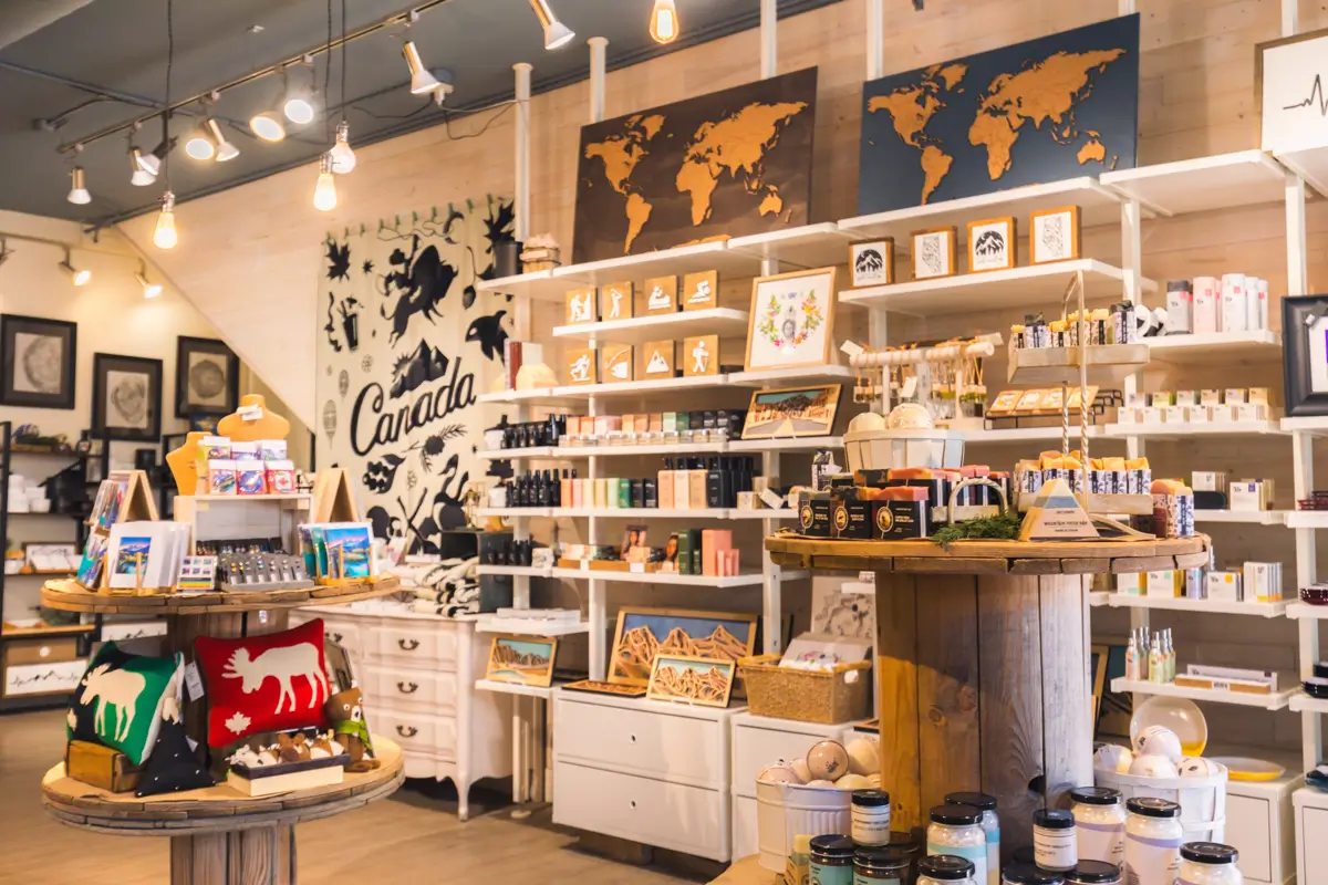 A boutique store interior in Canmore with shelves displaying various gift items, maps, artwork, and souvenirs. The decor features a large Canada mural and world map art on the walls, while wooden tables showcase additional products.