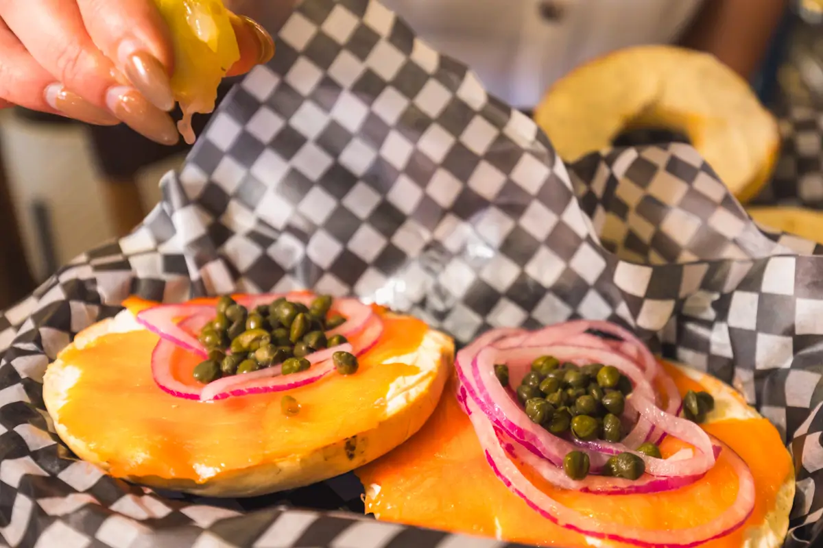 A close-up of two bagel halves topped with smoked salmon, sliced red onions, and capers on checkered paper in a cozy Canmore café. A hand squeezes a lemon wedge over the bagels, adding a touch of freshness.