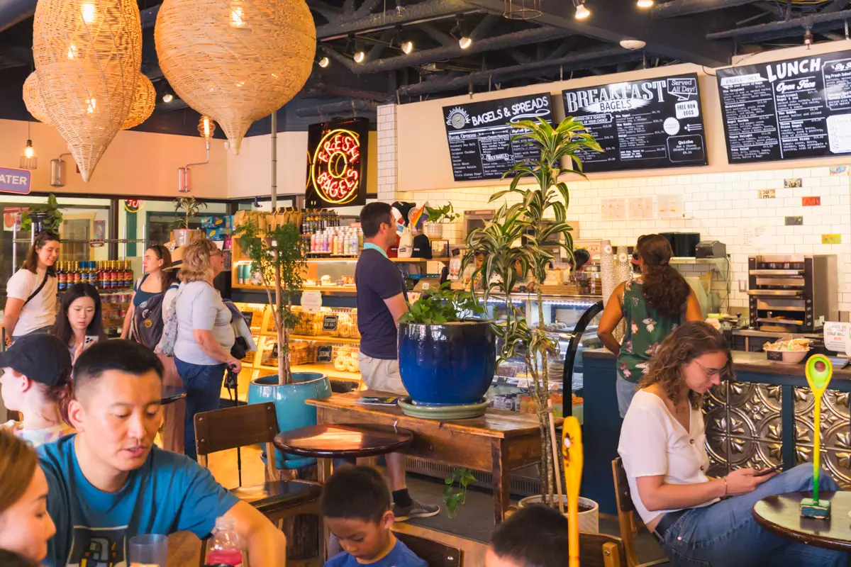 People are sitting and standing inside a busy Canmore café filled with plants, hanging lights, and a menu board on the wall. Some customers are ordering at the counter, while others eat or use their phones at tables.