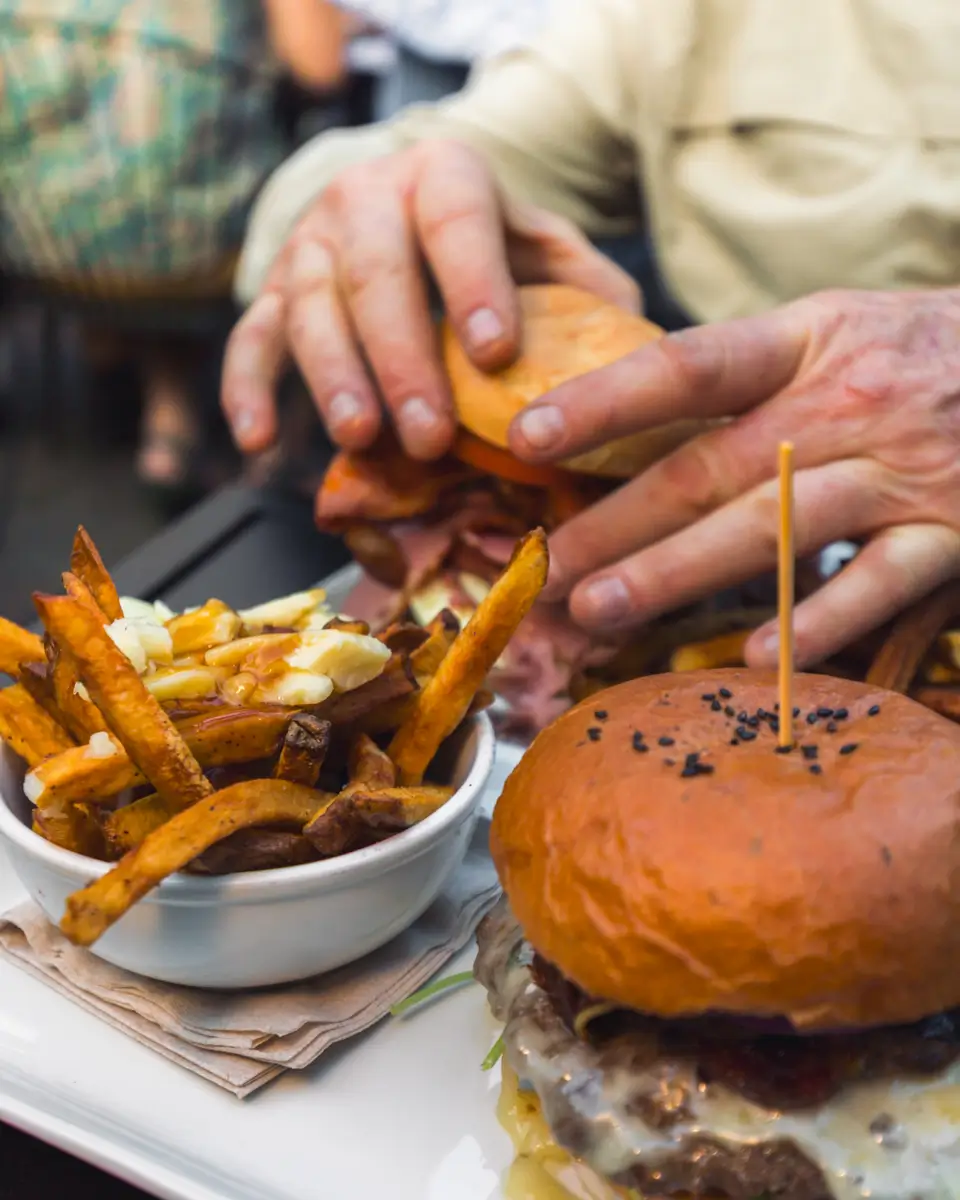 A close-up of a plate with a cheeseburger topped with a wooden skewer, a bowl of fries, and a person holding another sandwich in the background. The scene appears to be at a cozy restaurant in Canmore.