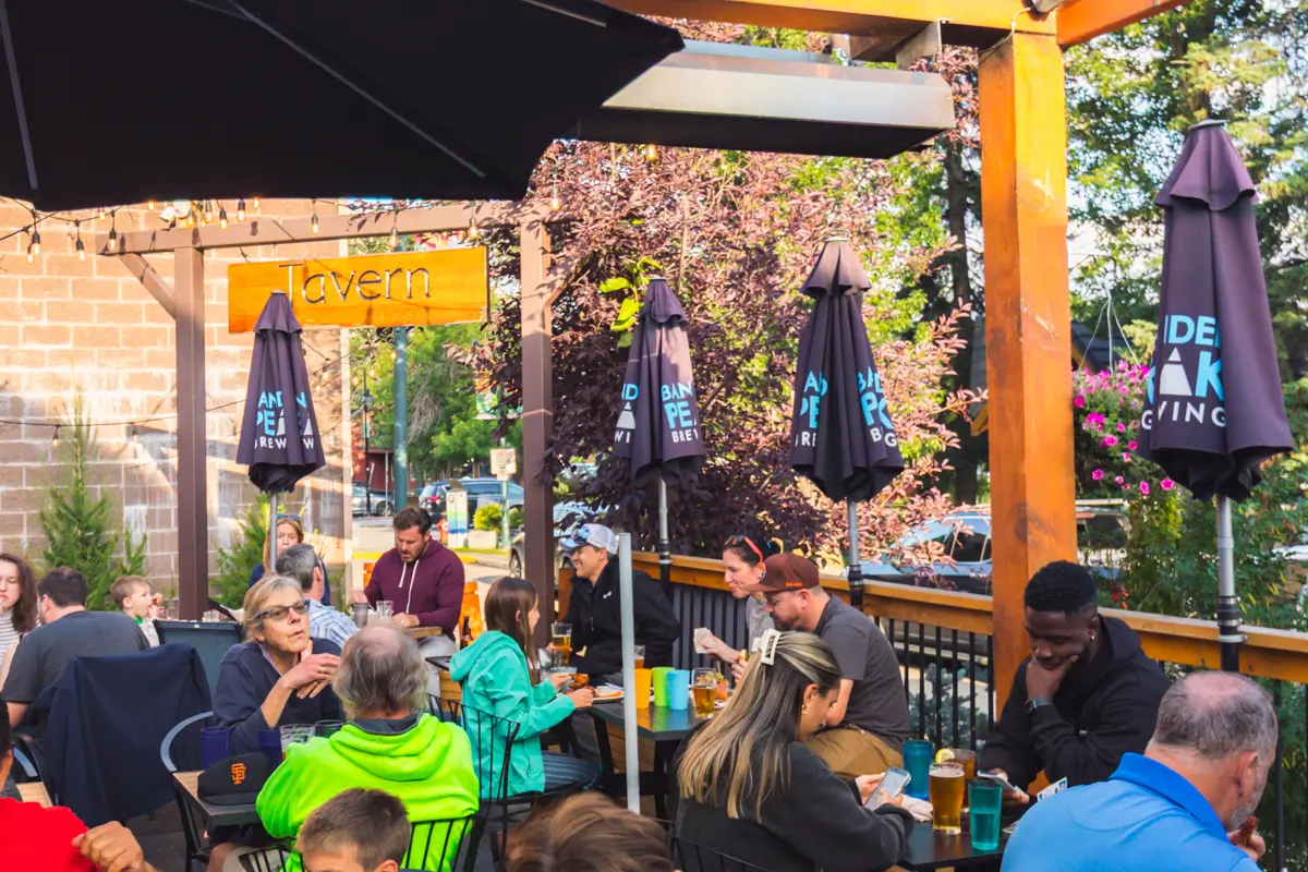 People sit at outdoor tables under umbrellas on a Canmore tavern patio, eating, drinking, and talking. Drinks and food fill the tables, while trees and plants surround the inviting seating area.