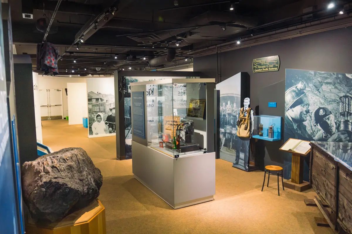 A Canmore museum exhibit displays mining artifacts in glass cases, with informational panels, historic photos on the walls, a large rock sample, and various mining equipment throughout the room.