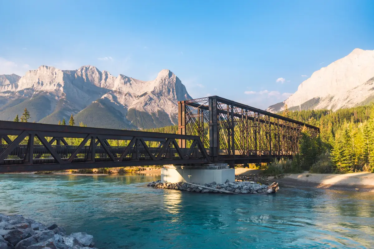 A steel truss bridge crosses a turquoise river with forested banks in Canmore, set against a backdrop of rugged mountains under a clear blue sky.
