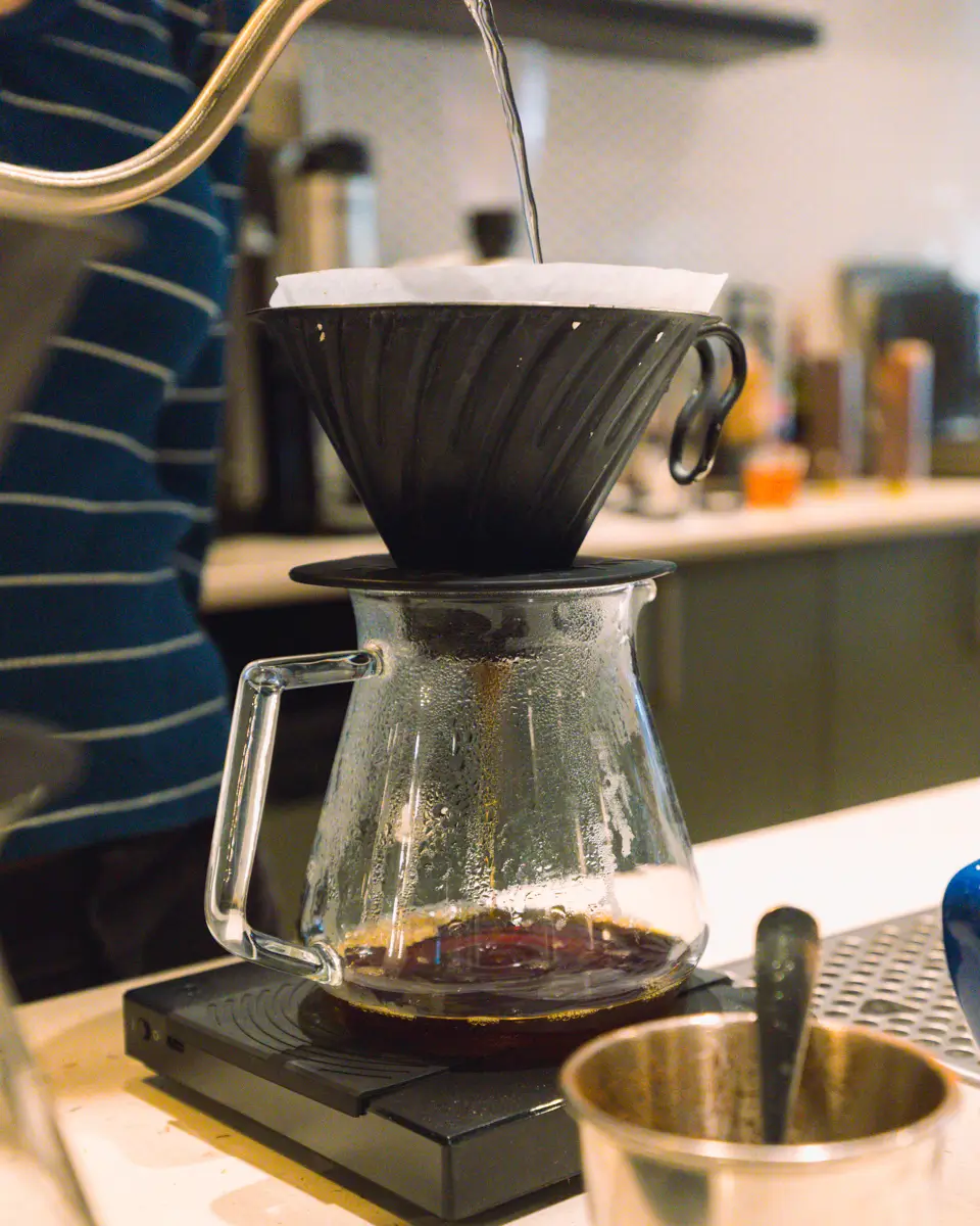 In Canmore, a person pours hot water from a kettle into a black pour-over coffee dripper with a paper filter, which sits atop a glass carafe placed on a digital scale.
