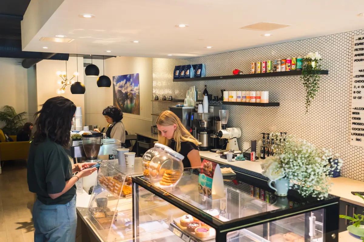 A cozy Canmore coffee shop interior with a display case of pastries, a barista preparing drinks behind the counter, and customers ordering. Shelves with coffee products and a menu board line the back wall.