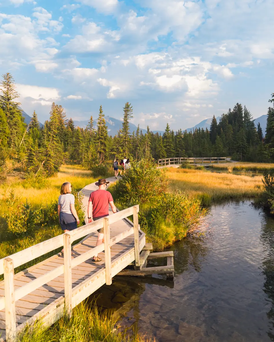 Several people walk on a wooden footbridge and pathway over a clear stream in a grassy, forested area near Canmore, with mountains and a partly cloudy sky in the background.