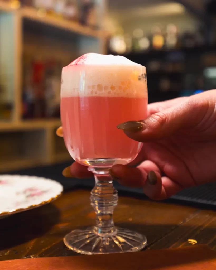 A hand with long, painted nails holds a glass of pink foamy cocktail on a wooden surface, set against blurred shelves and bottles in a cozy Canmore bar.