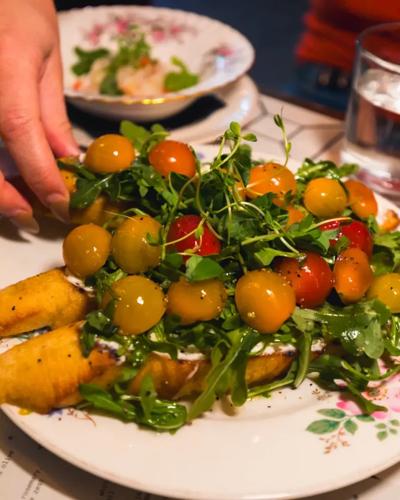 A hand reaches for a slice of toasted bread topped with cream, yellow and red cherry tomatoes, arugula, and microgreens on a floral-patterned plate—perfect for a cozy breakfast in Canmore. A glass of water and another dish appear in the background.