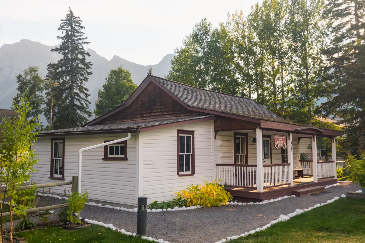 A small white wooden house with a covered front porch sits among trees in Canmore, with mountains visible in the background. A gravel path leads to the entrance, surrounded by lush greenery.