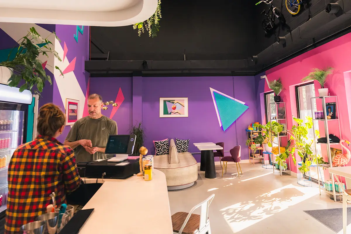 Two people interact at a counter in a brightly colored Canmore cafe with purple and pink walls, geometric decor, indoor plants, and sunlight streaming through large windows. Furniture and art add to the lively atmosphere.