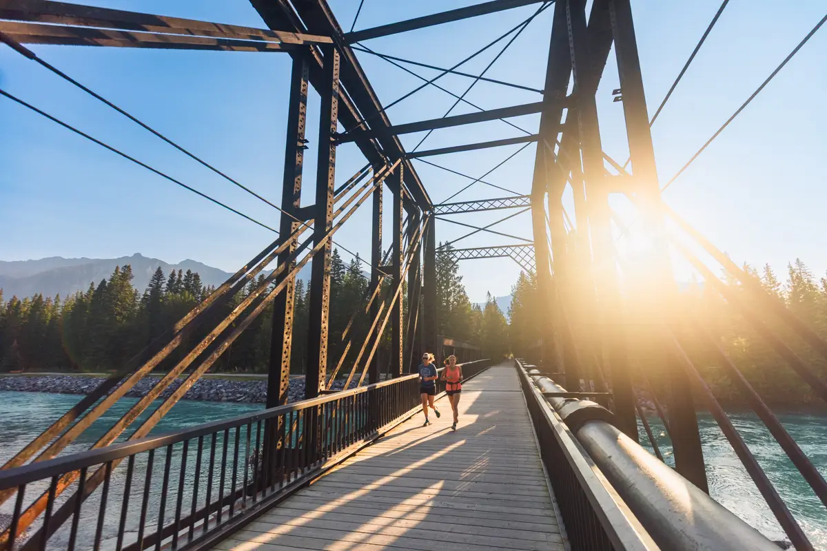 Two people jog across a metal truss bridge over a river in Canmore, surrounded by trees and mountains. Sunlight shines through the bridge structure, casting long shadows on the wooden walkway.