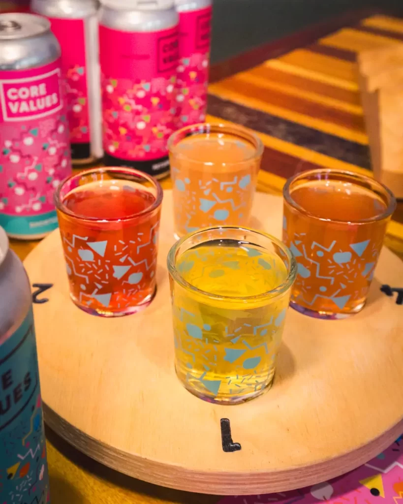 Four shot glasses filled with different colored liquids are arranged on a numbered wooden tray. Several cans labeled Core Values, inspired by the spirit of Canmore, are visible in the background.