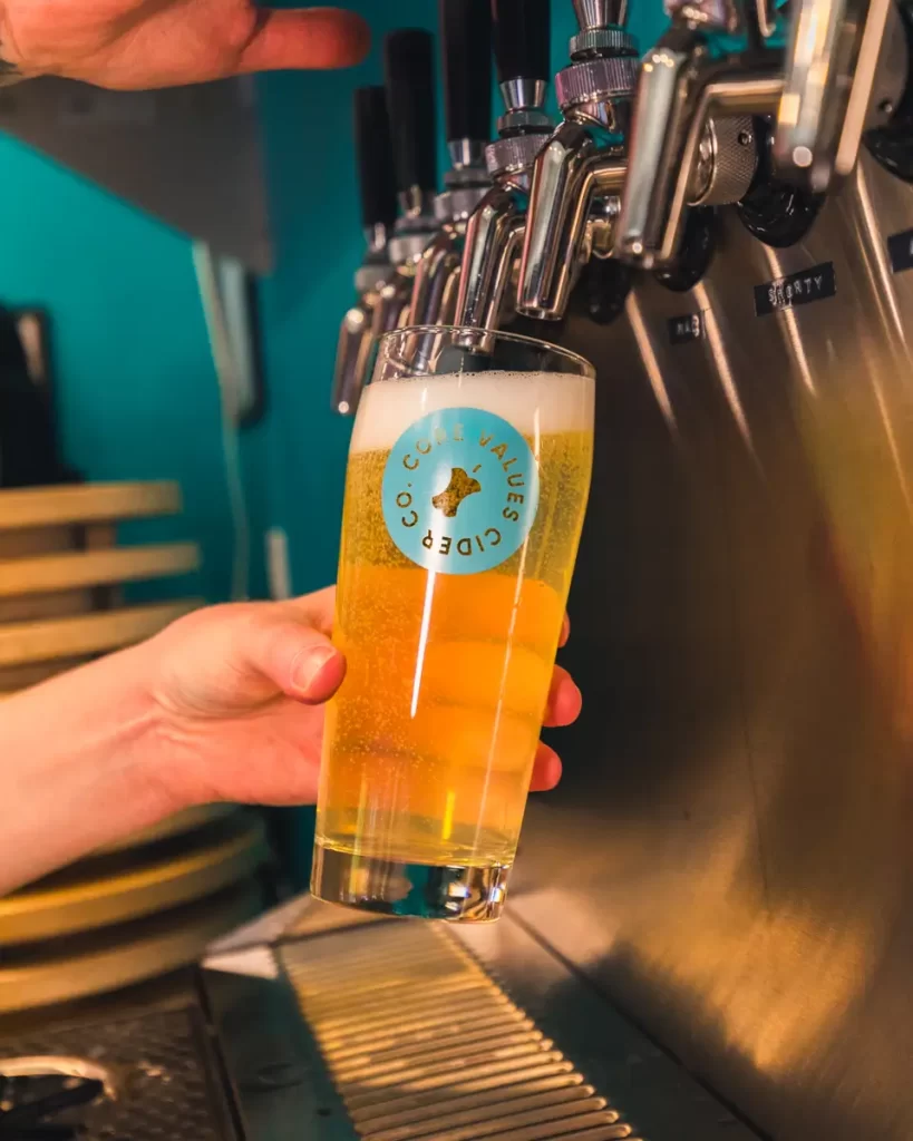 A hand fills a pint glass with light yellow beer from a tap at a Canmore bar. The glass, labeled “Core Values Cider,” has a foamy head, with several taps visible in the background.