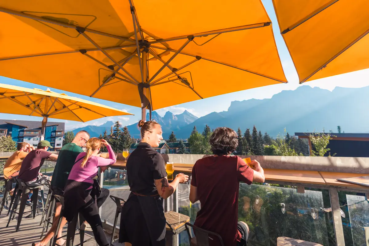 People sit at an outdoor patio in Canmore under large yellow umbrellas, enjoying mountain scenery in the background. Some are talking or holding drinks, and the weather appears sunny and clear.