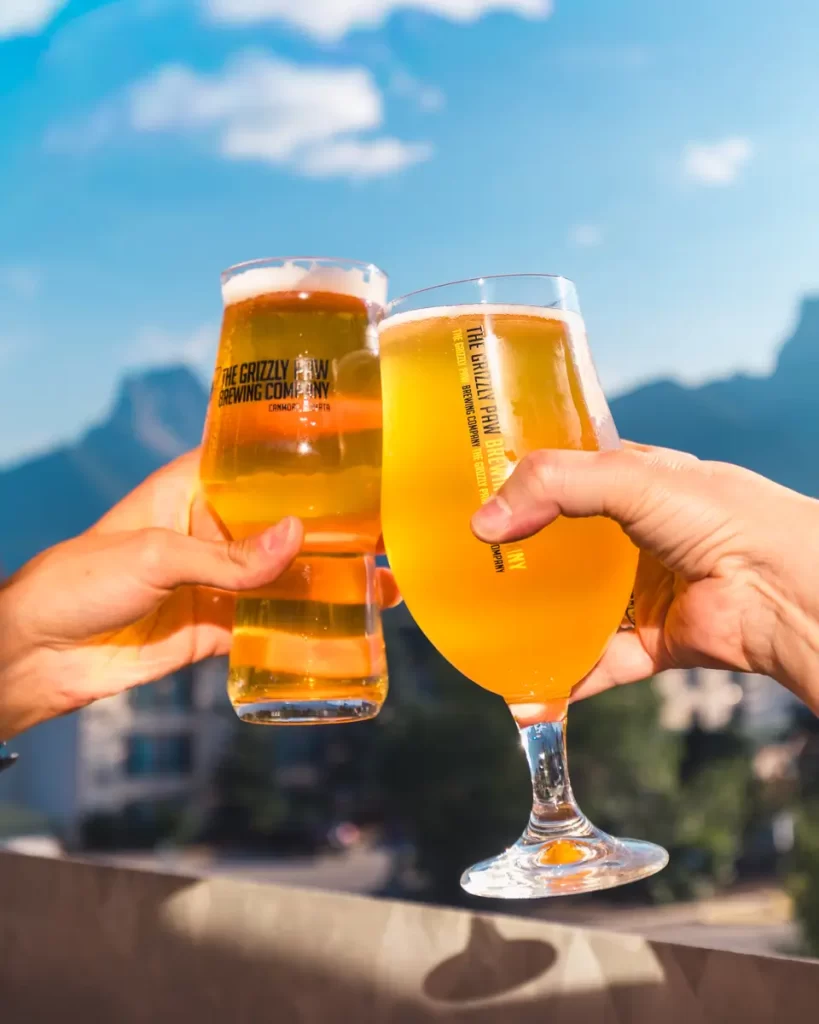 Two hands clink glasses of beer outdoors in Canmore, with a clear blue sky and blurred mountains in the background. One glass is a pint; the other is a stemmed beer glass, both featuring brewery logos.
