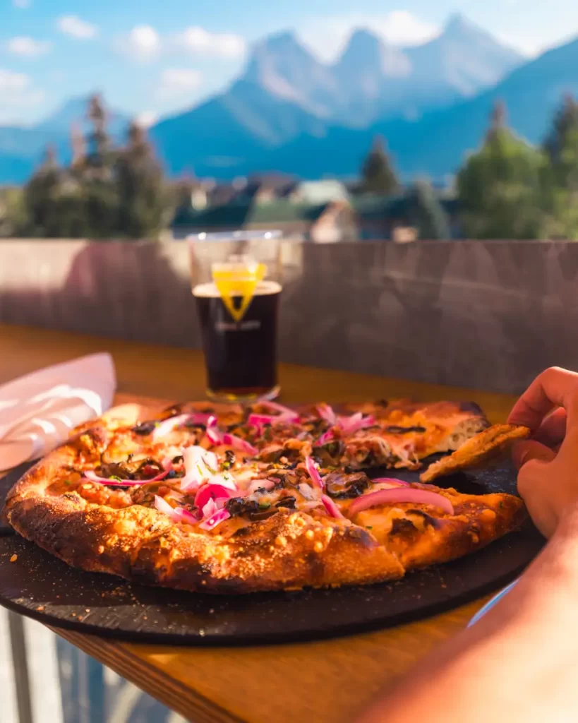A hand takes a slice of pizza topped with red onions and mushrooms from a round tray on a wooden table in Canmore, with a glass of dark beer and mountains blurred in the background.