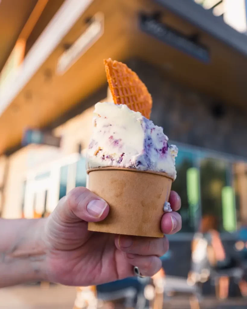 A hand holds a brown paper cup filled with two scoops of ice cream topped with a waffle piece. The scene is set outdoors in Canmore, with a building and blurred people sitting in the background.