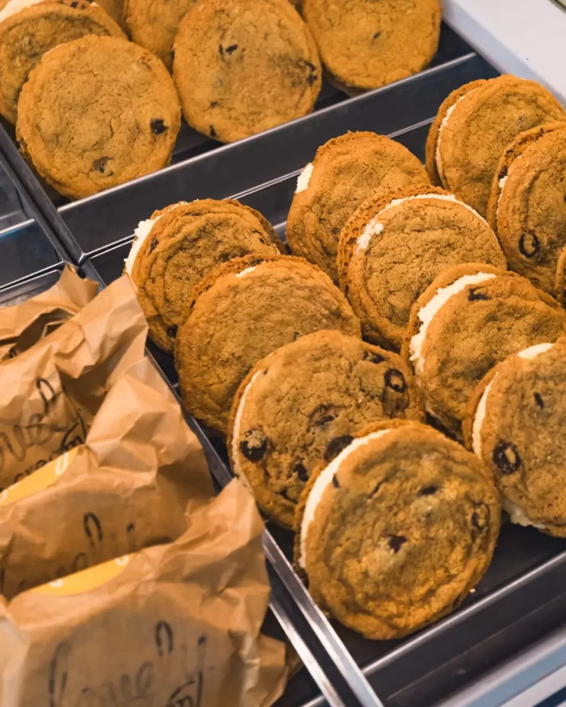 Rows of chocolate chip cookie ice cream sandwiches are arranged in metal trays in Canmore, with some wrapped in paper and others unwrapped, revealing the creamy ice cream filling between the cookies.