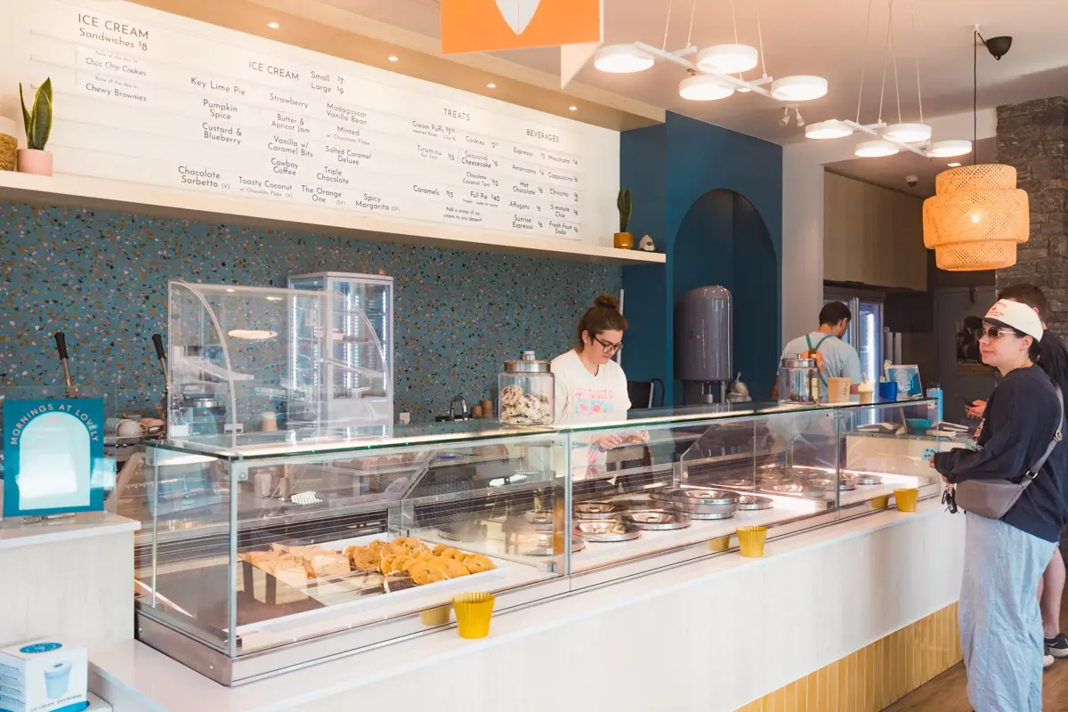 A modern ice cream shop interior in Canmore features a glass display case of pastries and tubs of ice cream. Two employees stand behind the counter, while a customer in sunglasses waits at the front. The menu is visible on the wall.