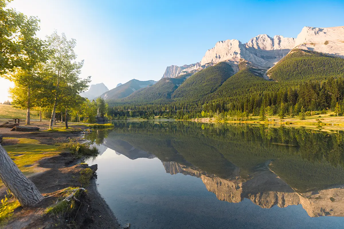 A calm lake near Canmore reflects trees, green forest, and rocky mountains under a clear blue sky. Sunlight illuminates the left side, with a picnic table by the water’s edge and a few leaning trees in the foreground.
