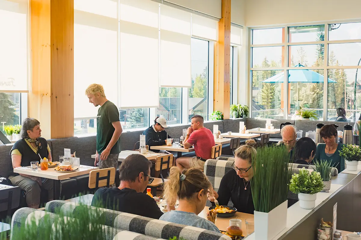 People sit and eat at tables in a bright, modern Canmore restaurant with large windows. A server stands by one table. Potted plants decorate the tables and dividers. Sunlight streams in, and trees are visible outside.