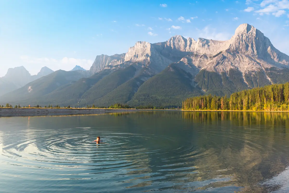 A person swims in a calm lake near Canmore, creating ripples in the water. Tall mountains rise in the background under a clear blue sky, with trees lining the edge of the lake.