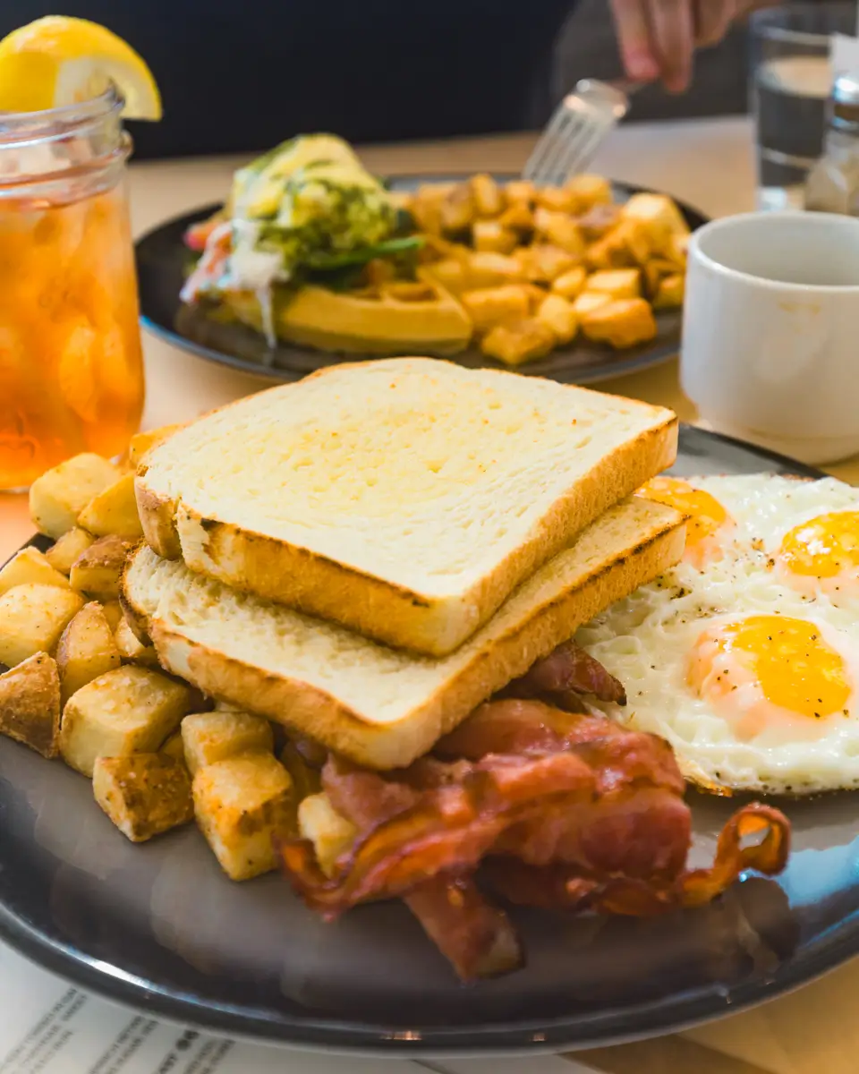 A plate with sunny-side-up eggs, crispy bacon, breakfast potatoes, and two slices of toasted bread. In the background are another plate of food, a glass of iced tea with lemon, and a white coffee mug in a cozy Canmore setting.
